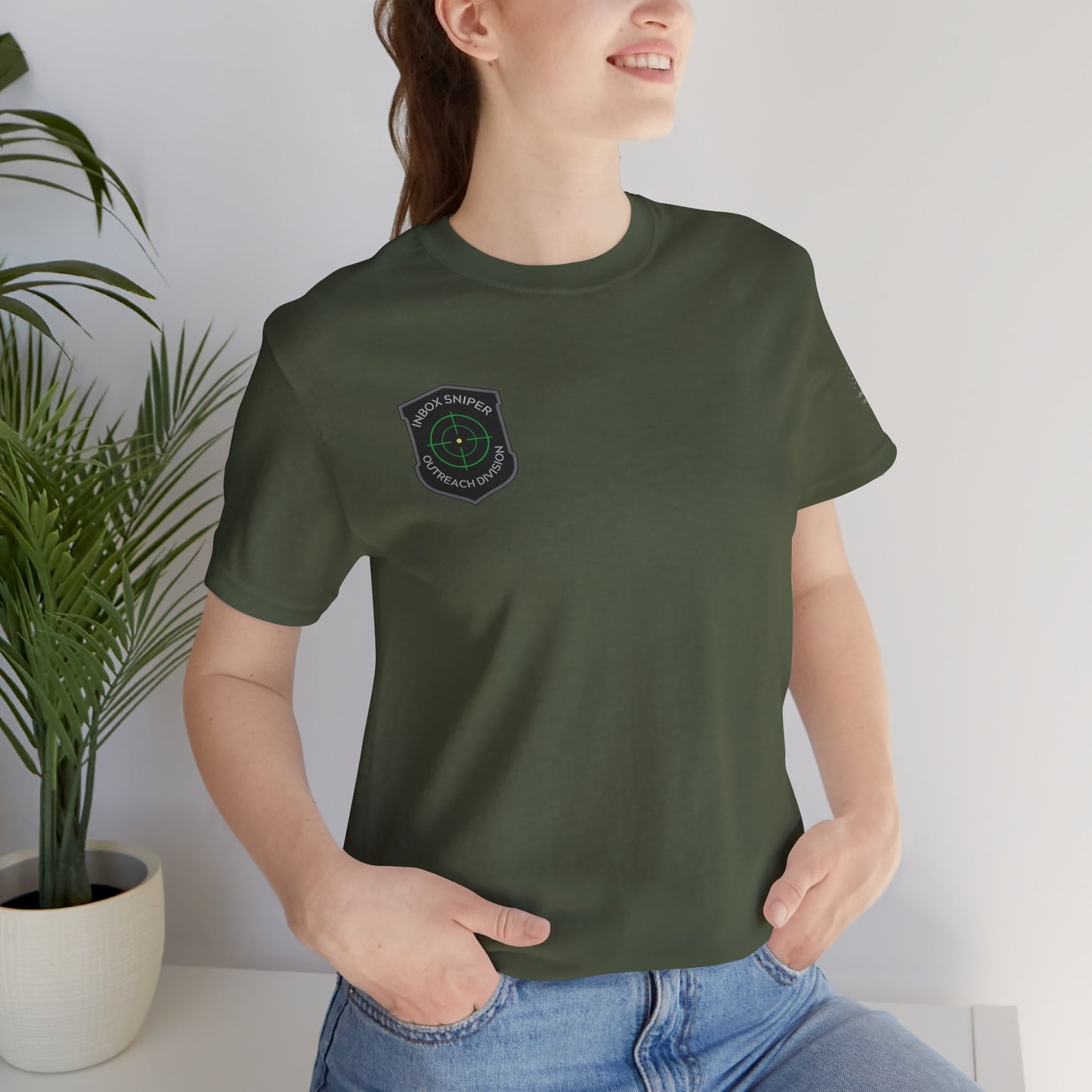 Person wearing a green t-shirt with a logo, standing indoors next to a plant.